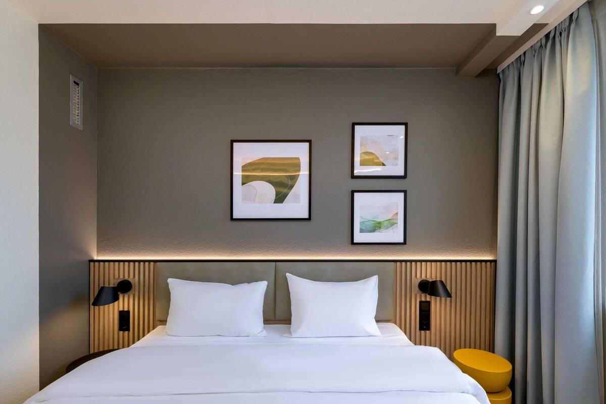 A hotel room with a bed, two wall-mounted lamps, and three framed artworks on the wall.