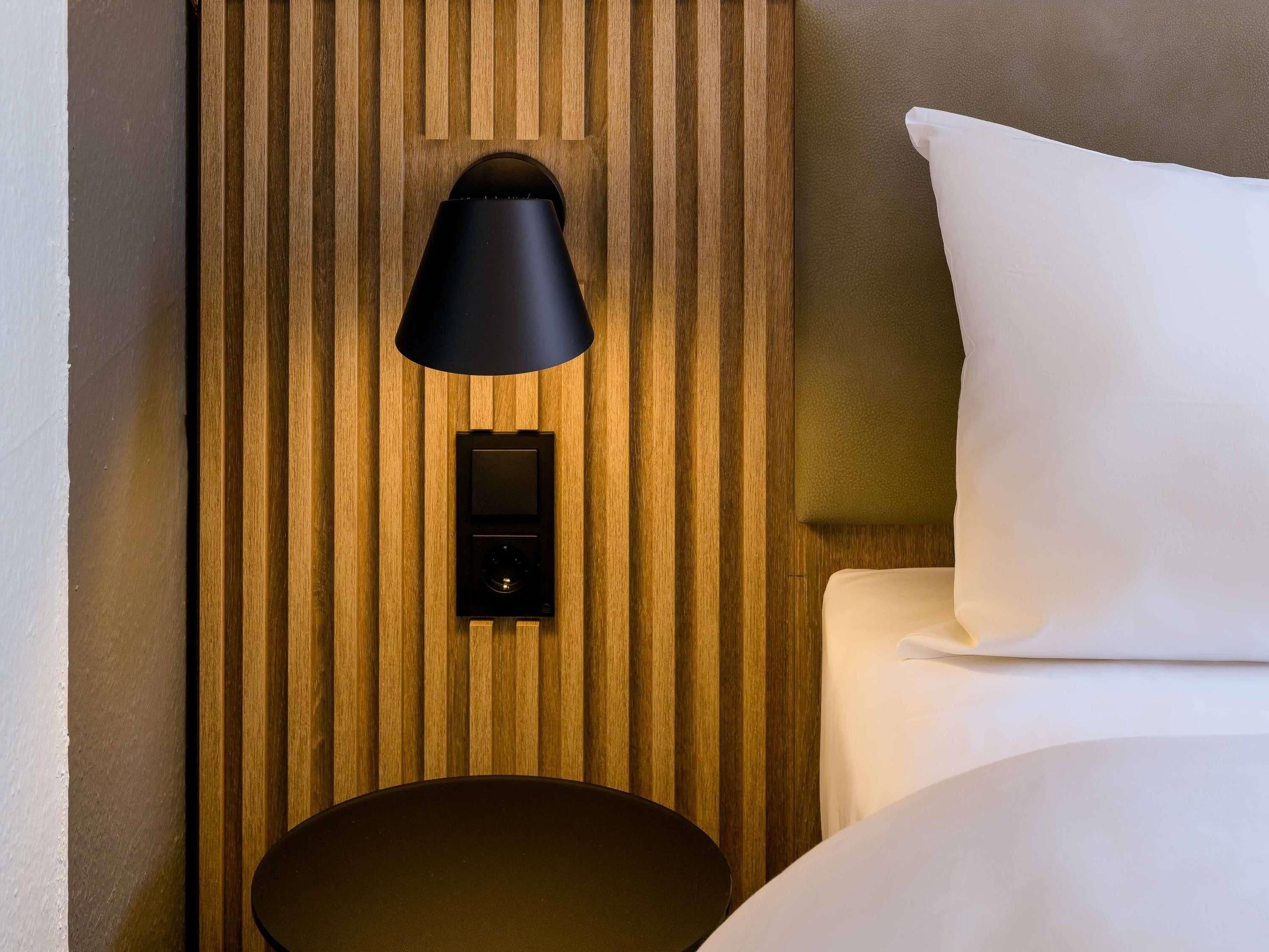 Une chambre d’hôtel avec un mur lambrissé en bois, une lampe noire fixée au mur, un panneau de commande et un oreiller blanc.