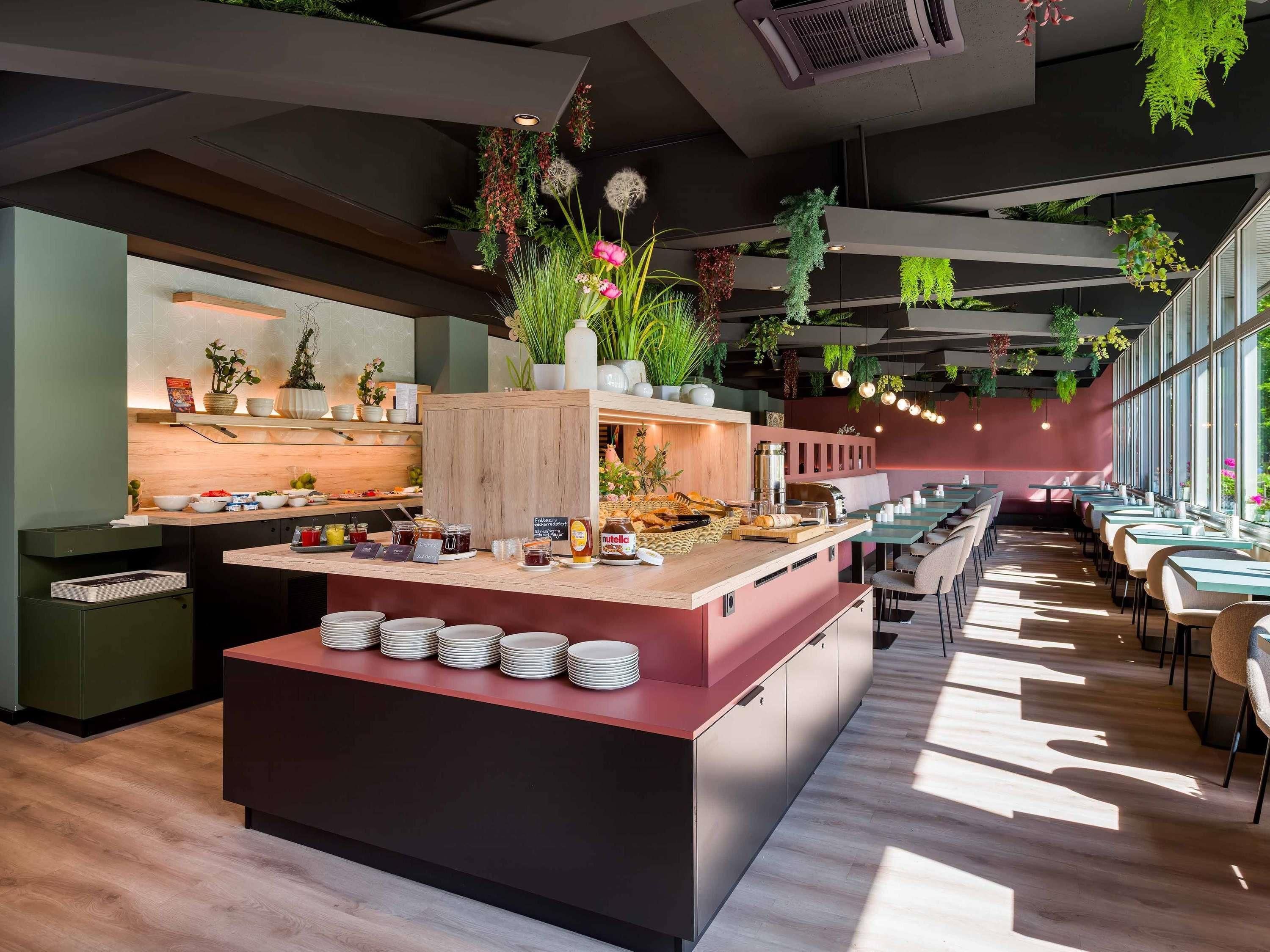 Ein modernes Restaurant mit einer zentralen Insellounge, Esstischen und hängenden Pflanzen.
