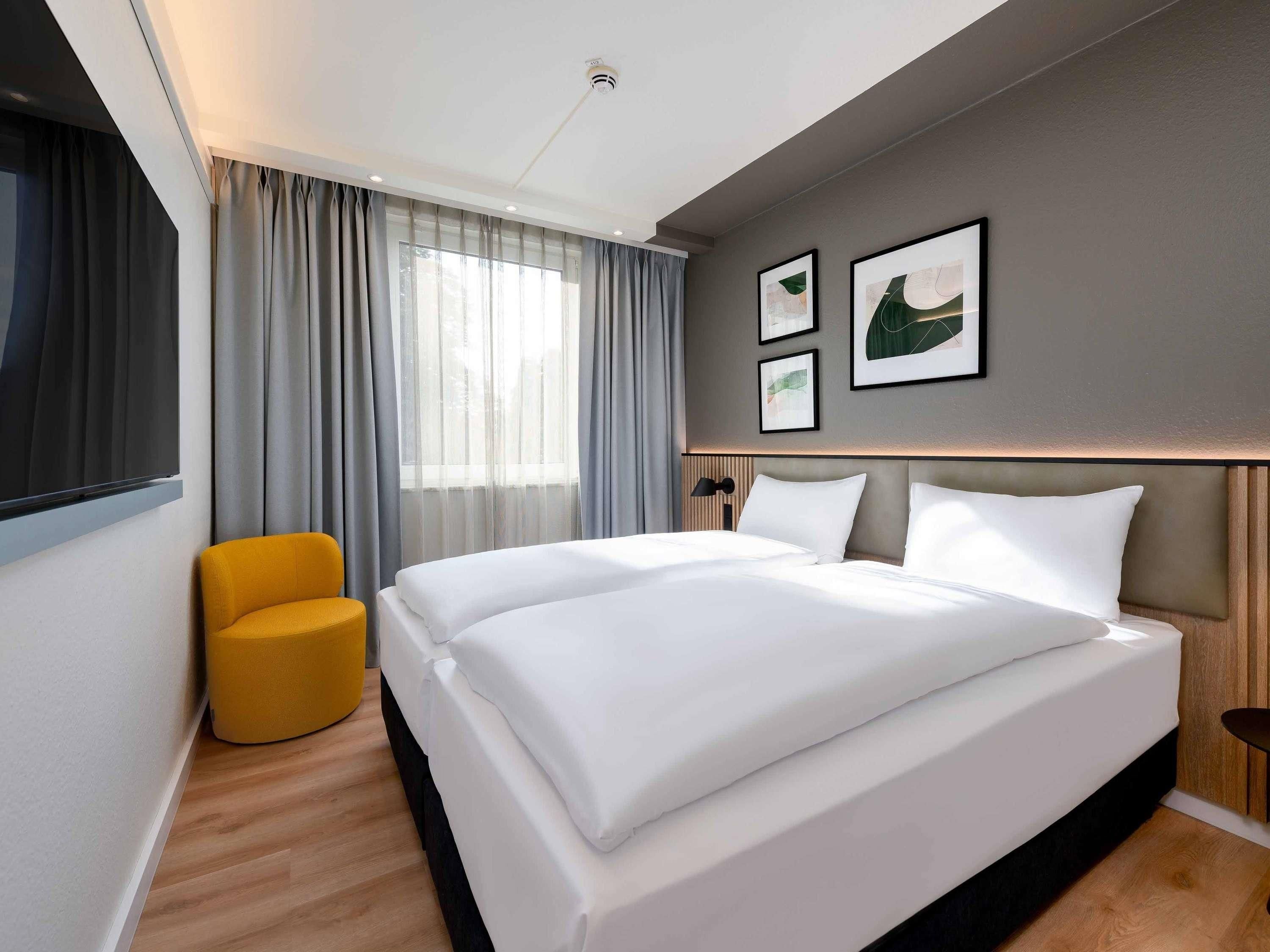 Une chambre d’hôtel avec un grand lit, un fauteuil jaune, une fenêtre avec des rideaux et des œuvres d’art encadrées accrochées au mur.
