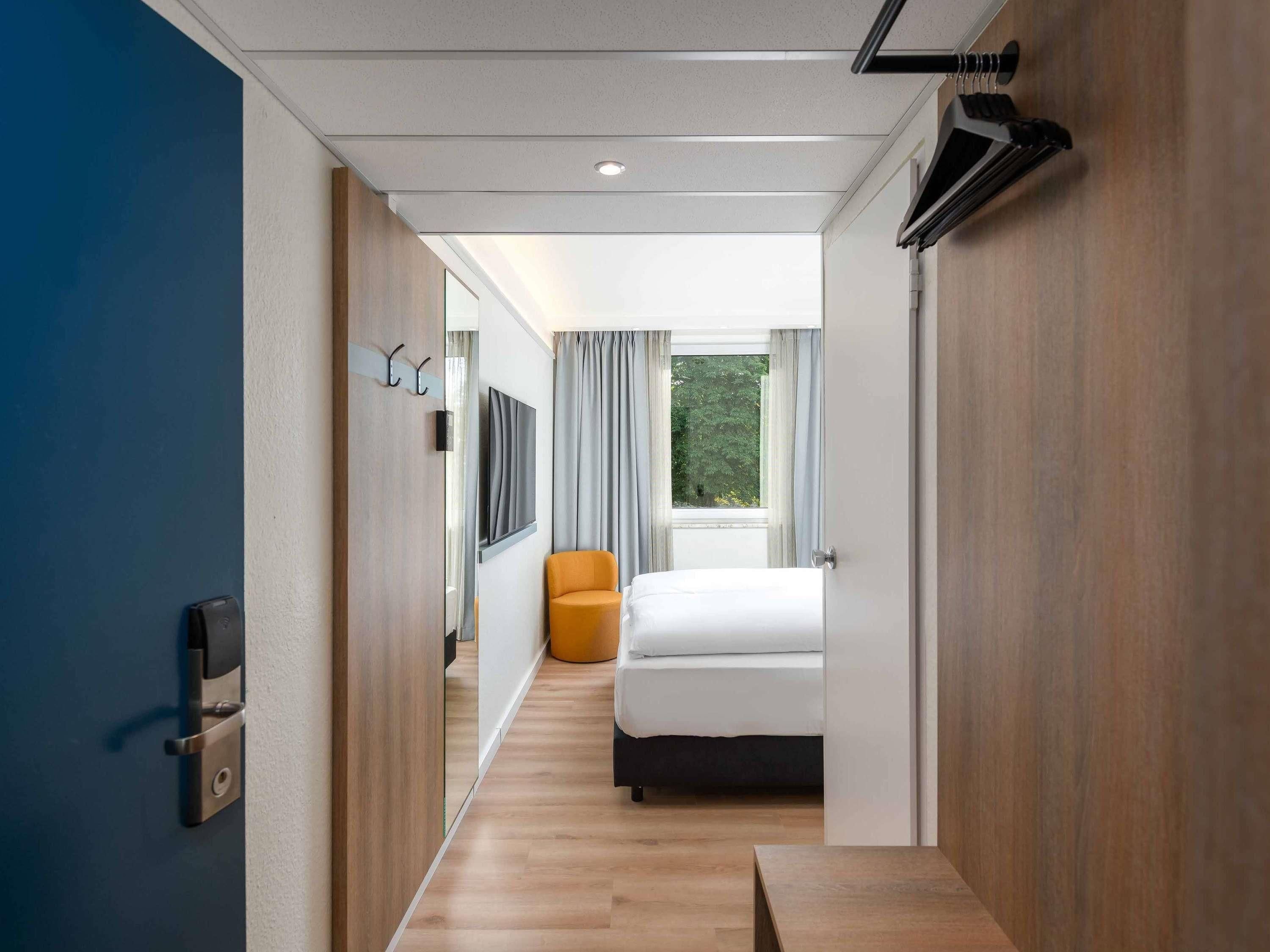 A modern hotel room with a bed, a chair, a television, and a window with curtains.