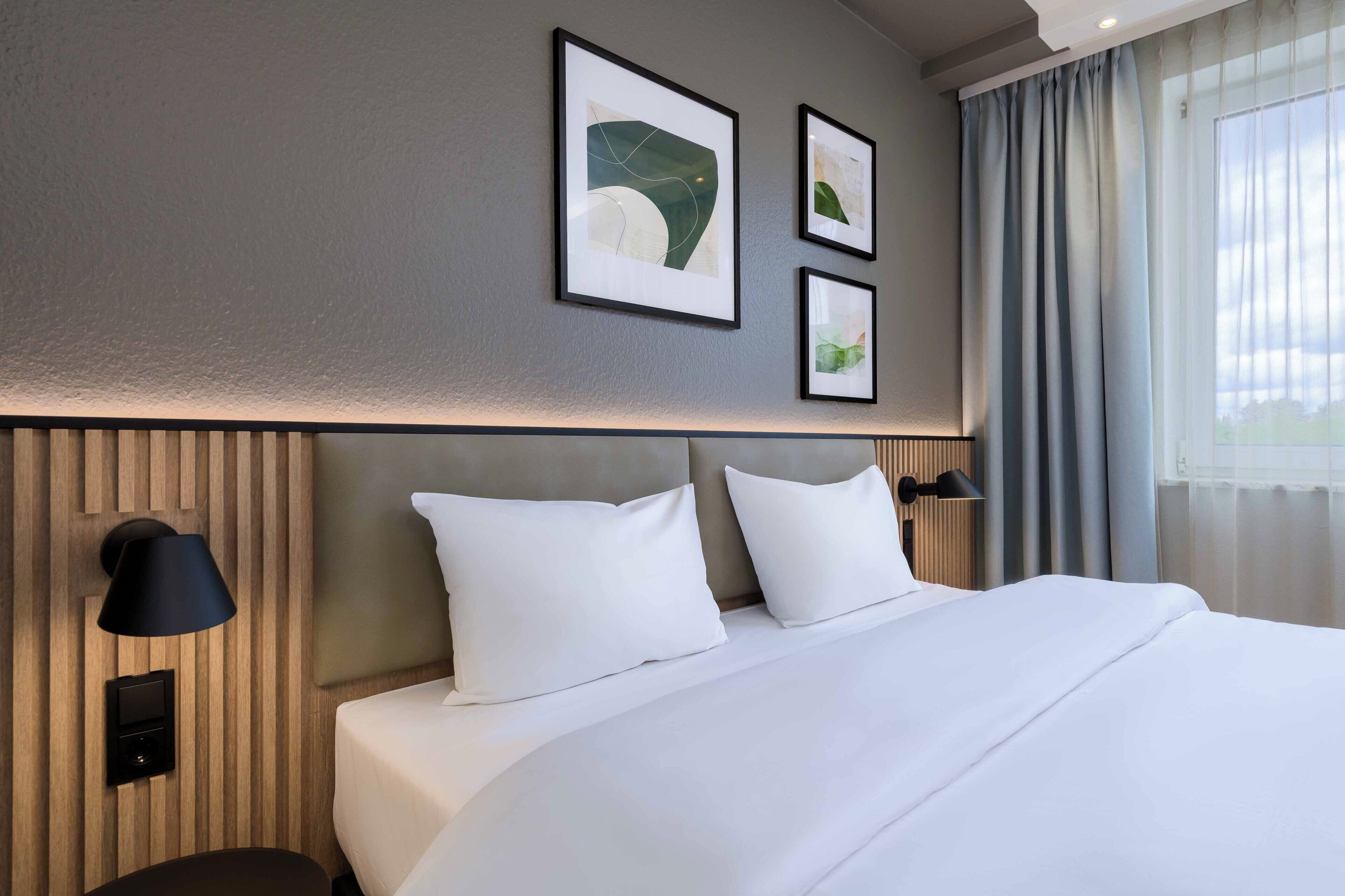 A hotel room with a bed, wall-mounted artwork, a lamp, and a window with curtains.