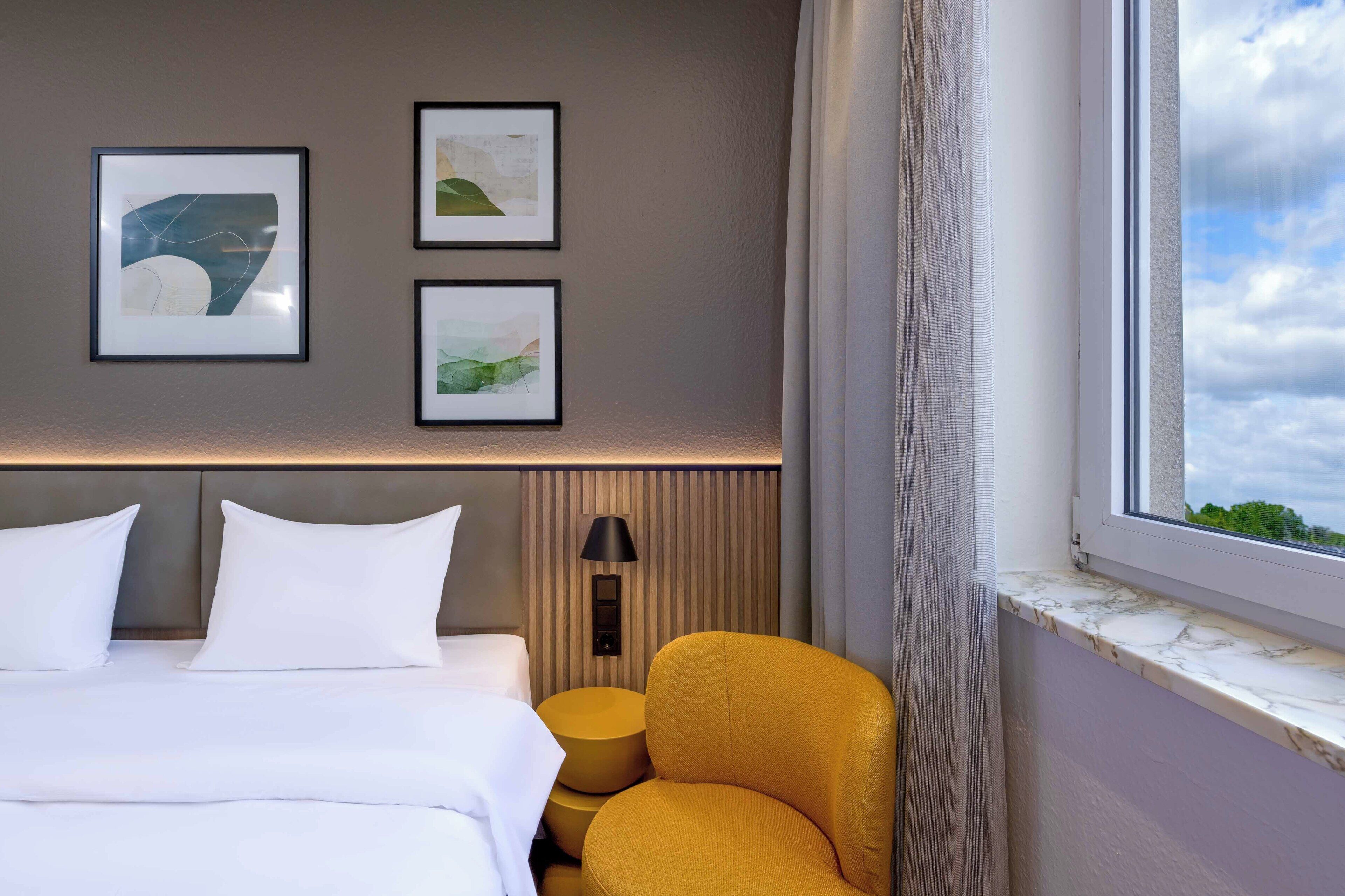 A hotel room with a bed, two pillows, a headboard, a wall-mounted lamp, a window with a view, and three framed pictures on the wall.