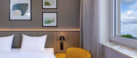 A hotel room with a bed, two pillows, a headboard, a wall-mounted lamp, a window with a view, and three framed pictures on the wall.
