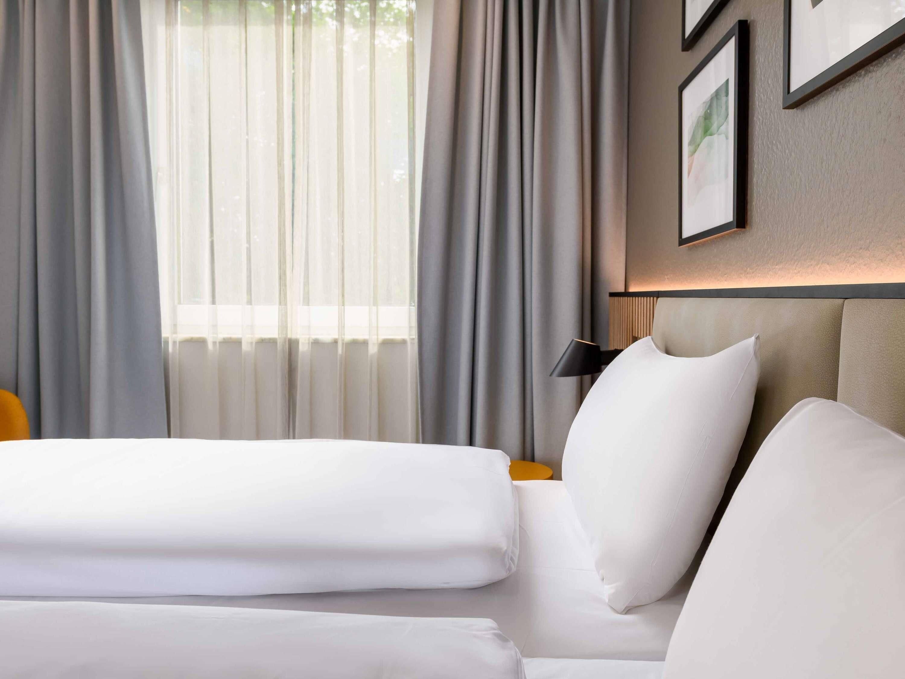 Habitació d'hotel amb llit, una tauleta de nit, una làmpada i una finestra amb cortines.