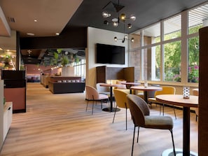 A modern interior with a wooden floor, seating area with round tables and chairs, a kitchen area with a counter, and large windows providing natural light. - ibis Paderborn City (Paderborn)