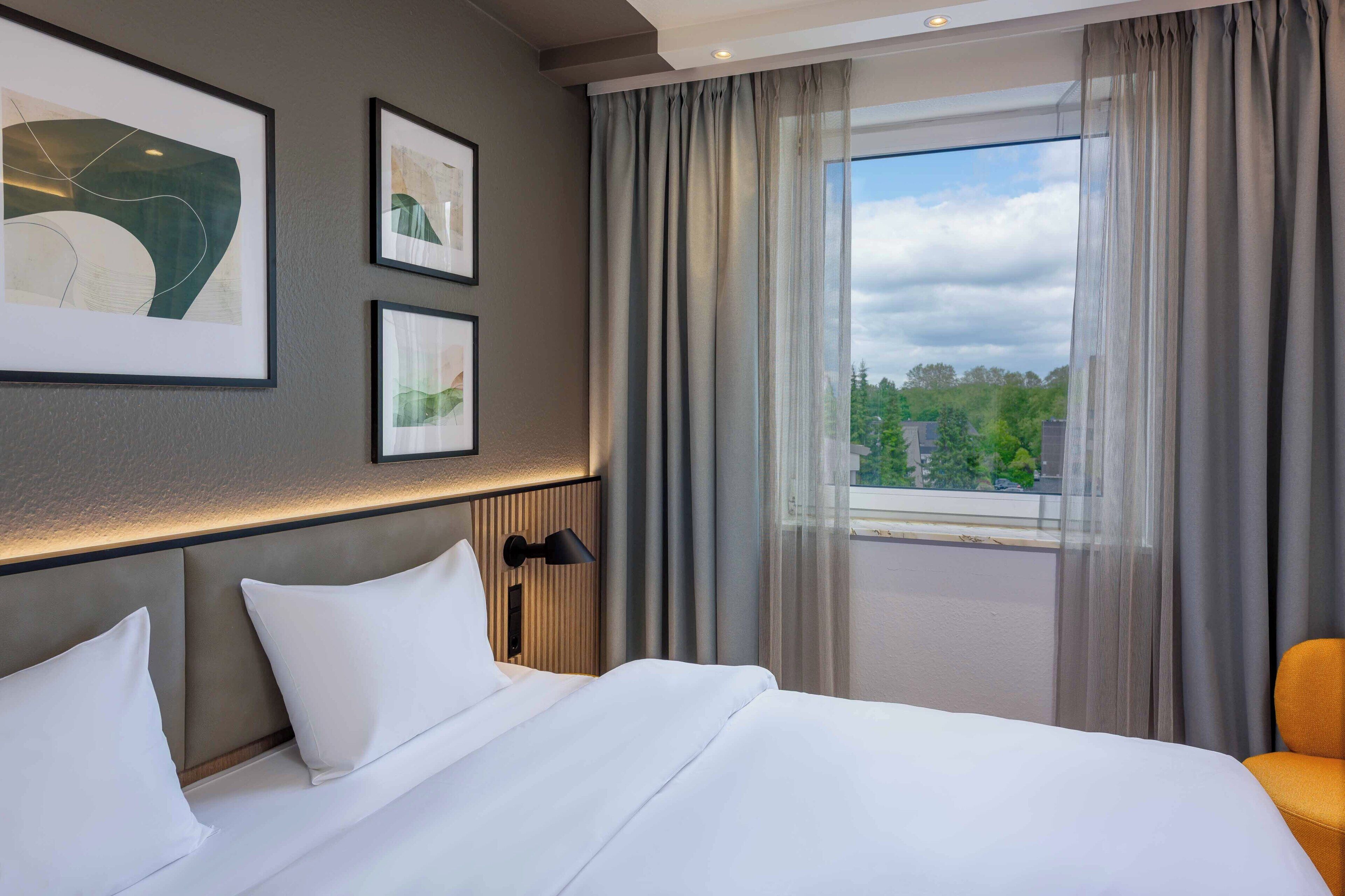 A hotel room with a bed, a nightstand, a lamp, and a window with curtains.