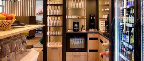 Zona de bar con estantes llenos de botellas y vasos, un refrigerador con bebidas y una barra con una cafetera.
