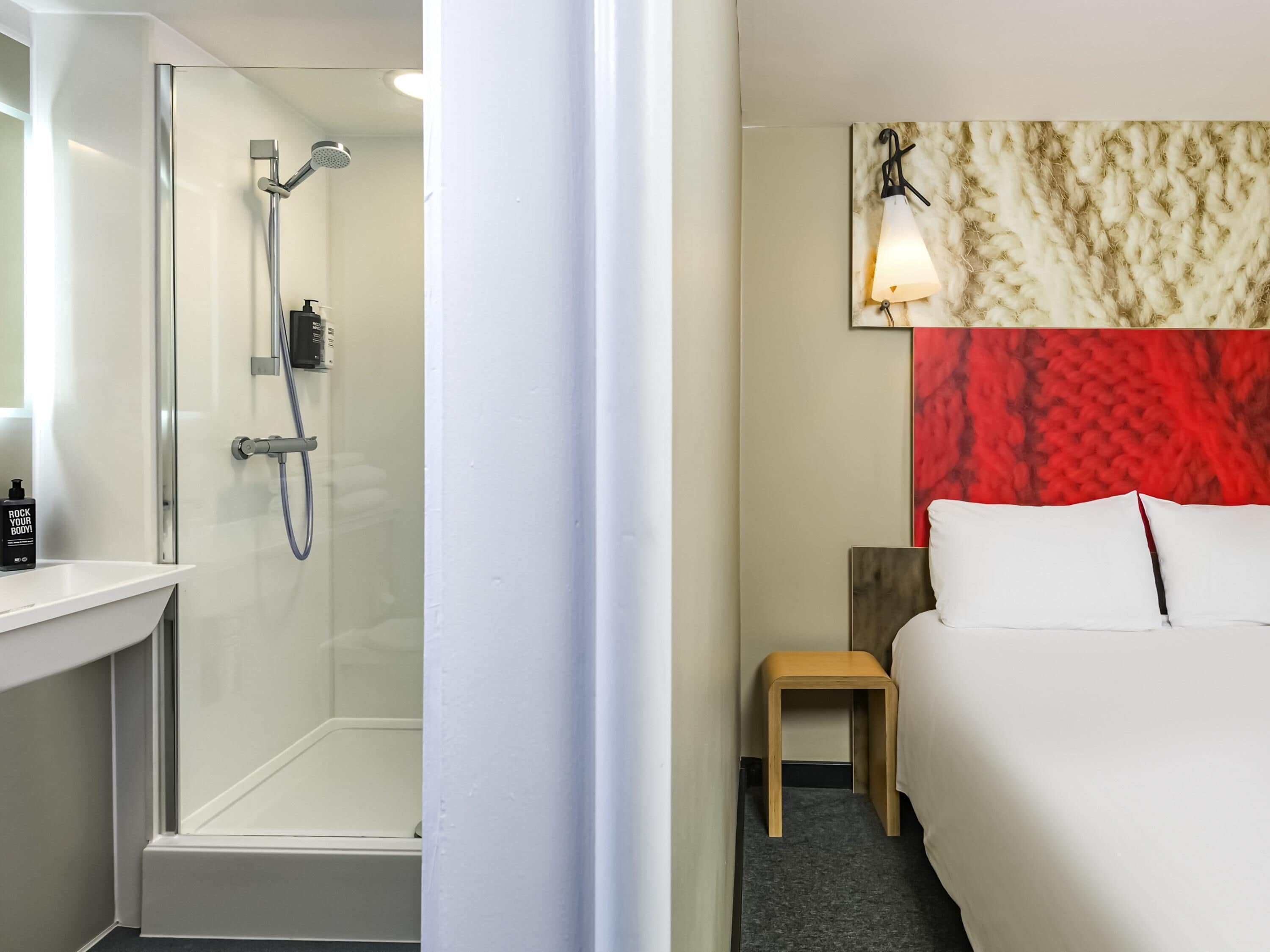 A hotel room with a shower, a bedside table, a bed, and a wall-mounted light fixture.