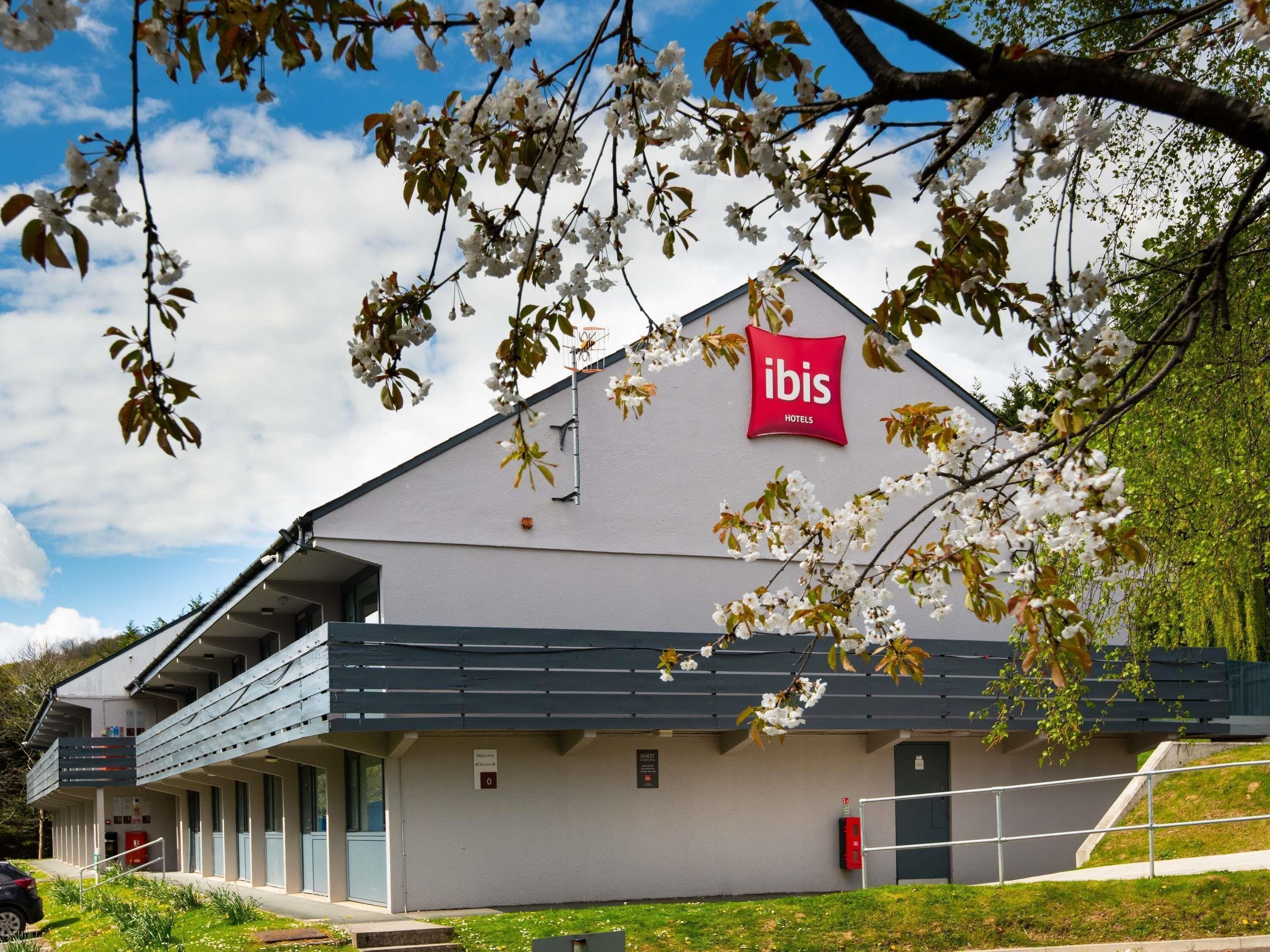 A modern hotel building with a prominent red logo, a covered outdoor area, and a sign indicating a guest entrance.