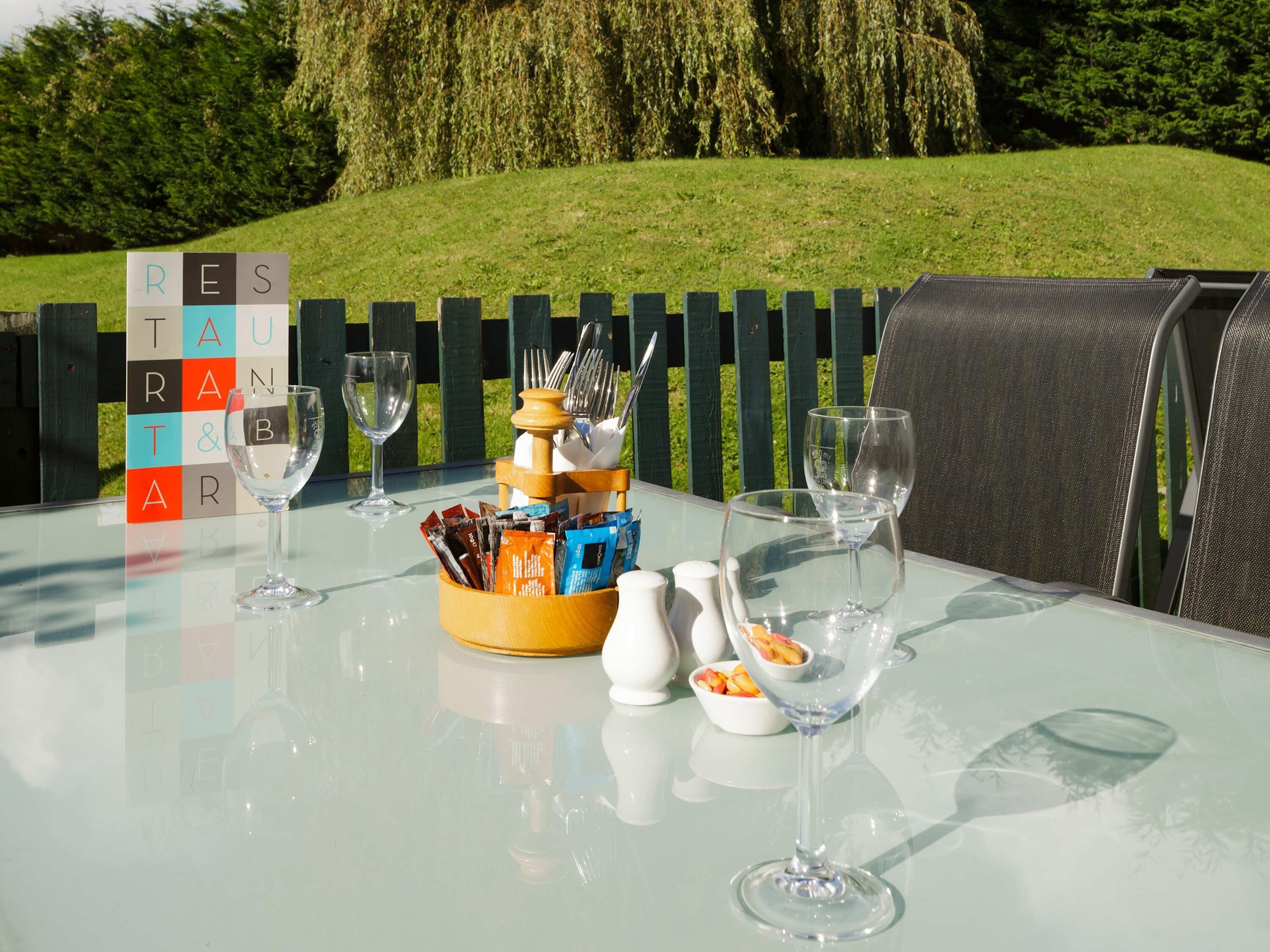 Mesa al aire libre con cubierta de vidrio, copas de vino, salero y pimentero, un cuenco de fruta y un bloque de madera con letras.