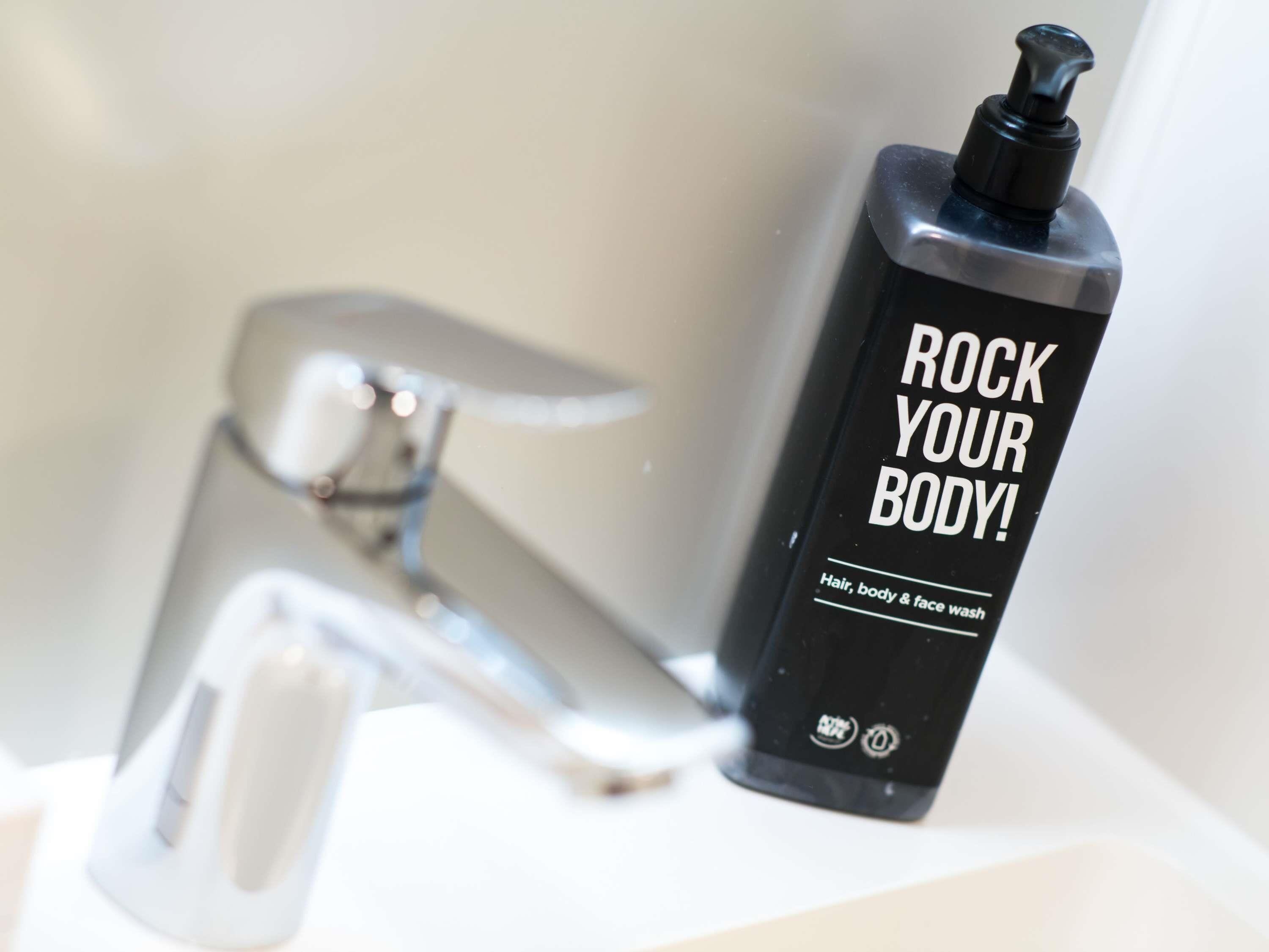 Een zwarte fles met een pompdispenser waarop staat 'ROCK YOUR BODY!' naast een chromen kraan.