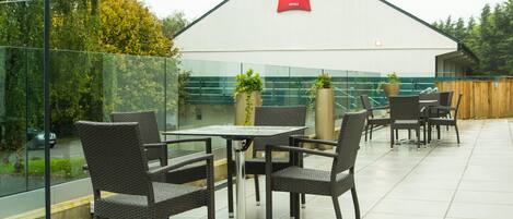 An outdoor seating area with wicker chairs and tables, a glass railing, and a view of trees and a building with an 'ibis Hotels' sign.