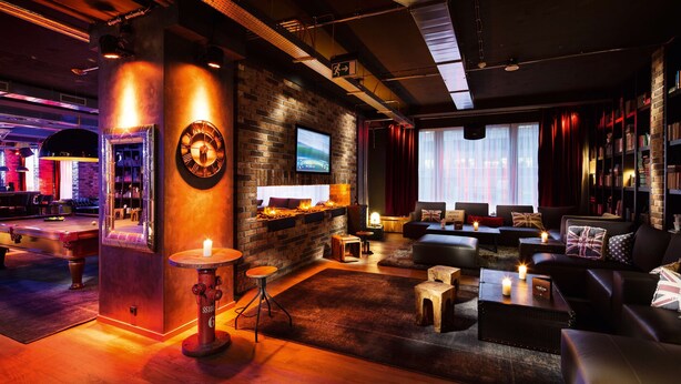 A cozy lounge area with a brick wall, a large clock, and a fireplace. There are multiple seating options including sofas and a small table with candles. - Hotel PULSE8 (Prague)