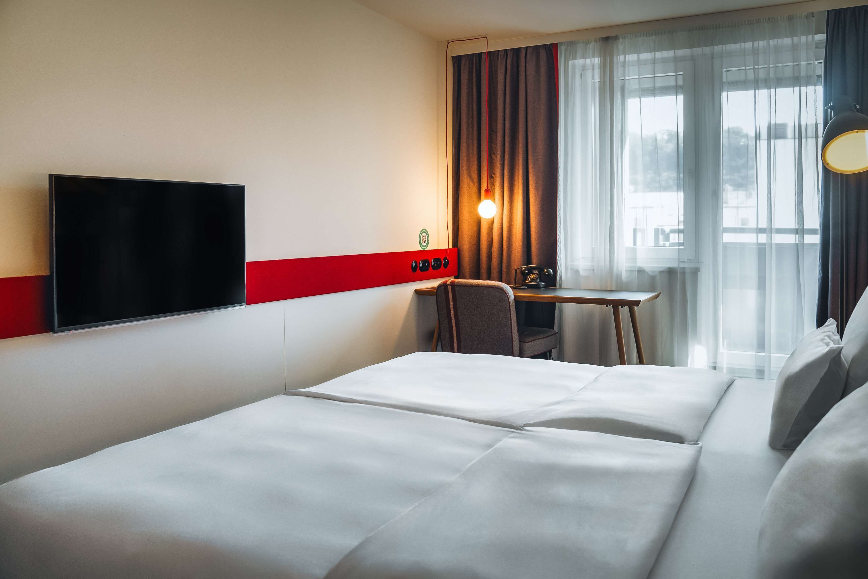 Ein Hotelzimmer mit einem großen Bett, einem Schreibtisch mit Stuhl, einem an der Wand montierten Fernseher und einem Fenster mit Vorhängen.