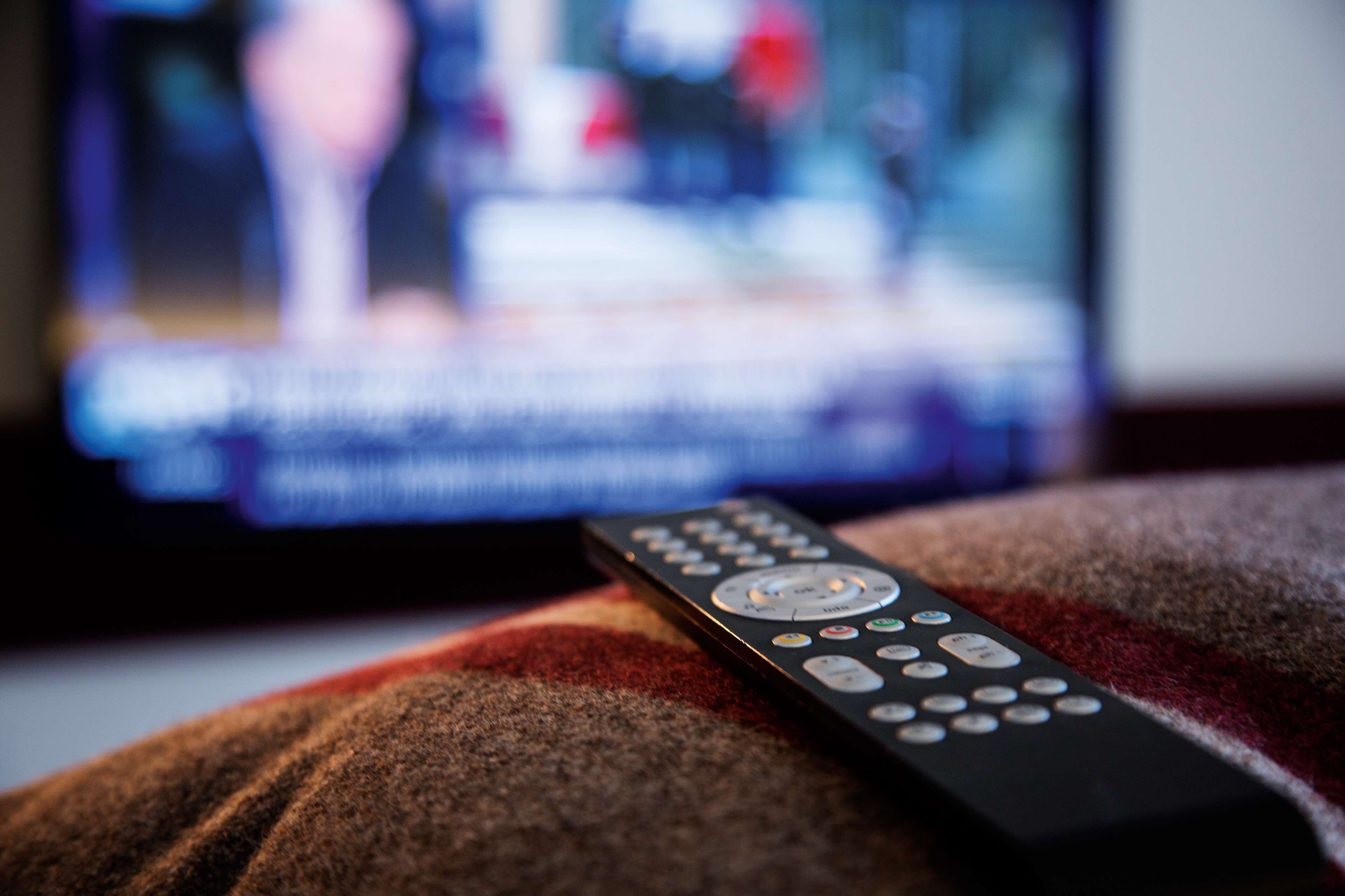 A remote control on a textured surface, likely a couch or chair, with a blurred television screen in the background.