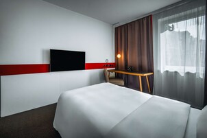 Quarto de hotel com cama, escrivaninha, cadeira, televisão na parede e janela com cortinas.