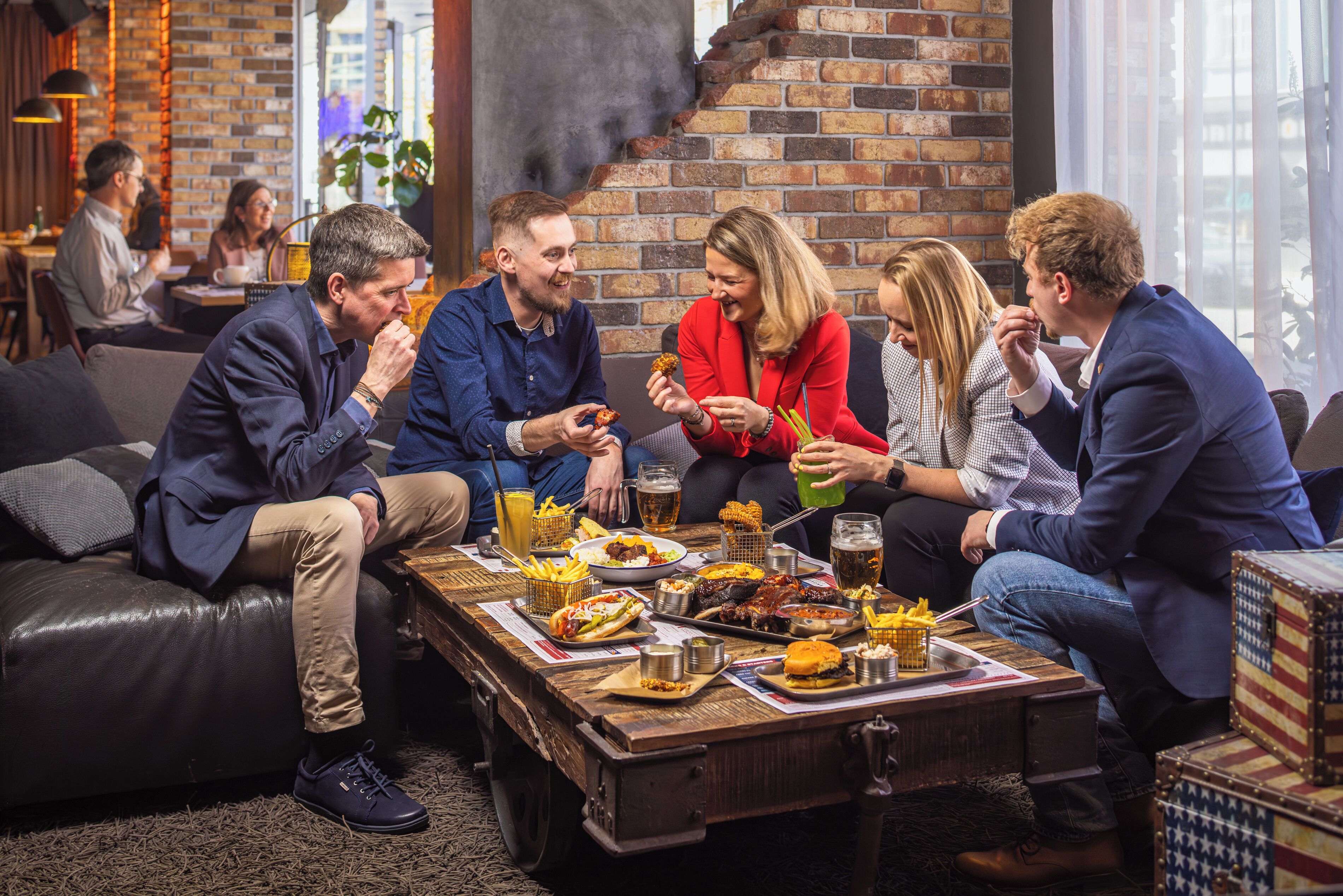 A group of people dining at a rustic wooden table with various dishes and drinks, in a cozy indoor setting with brick walls and leather seating.