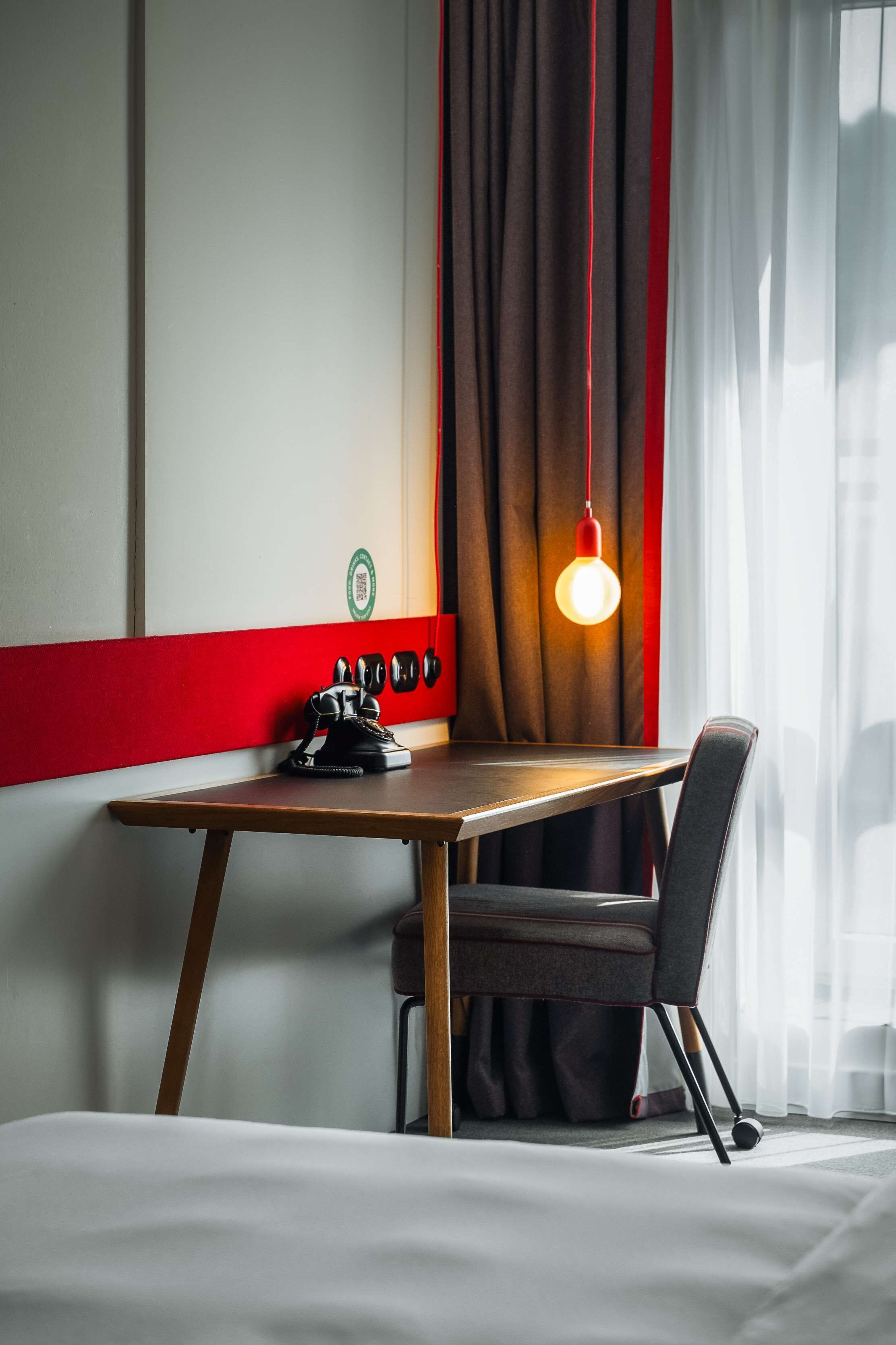 A hotel room with a wooden table, a chair, a desk lamp, and a window with curtains.