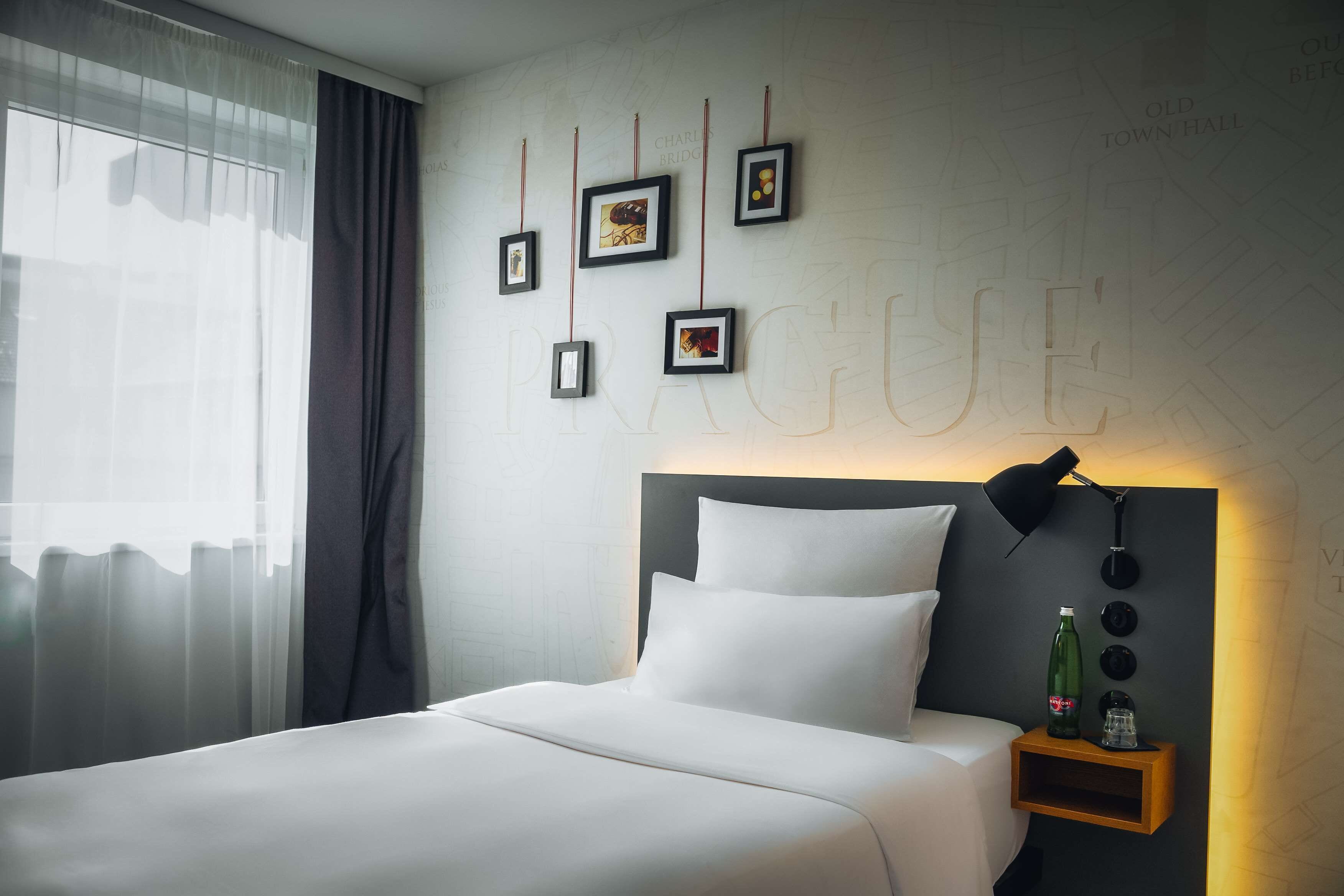 A modern hotel room with a neatly made bed, a wall-mounted picture frame, a bedside table with a bottle and glasses, and a large window with sheer curtains.