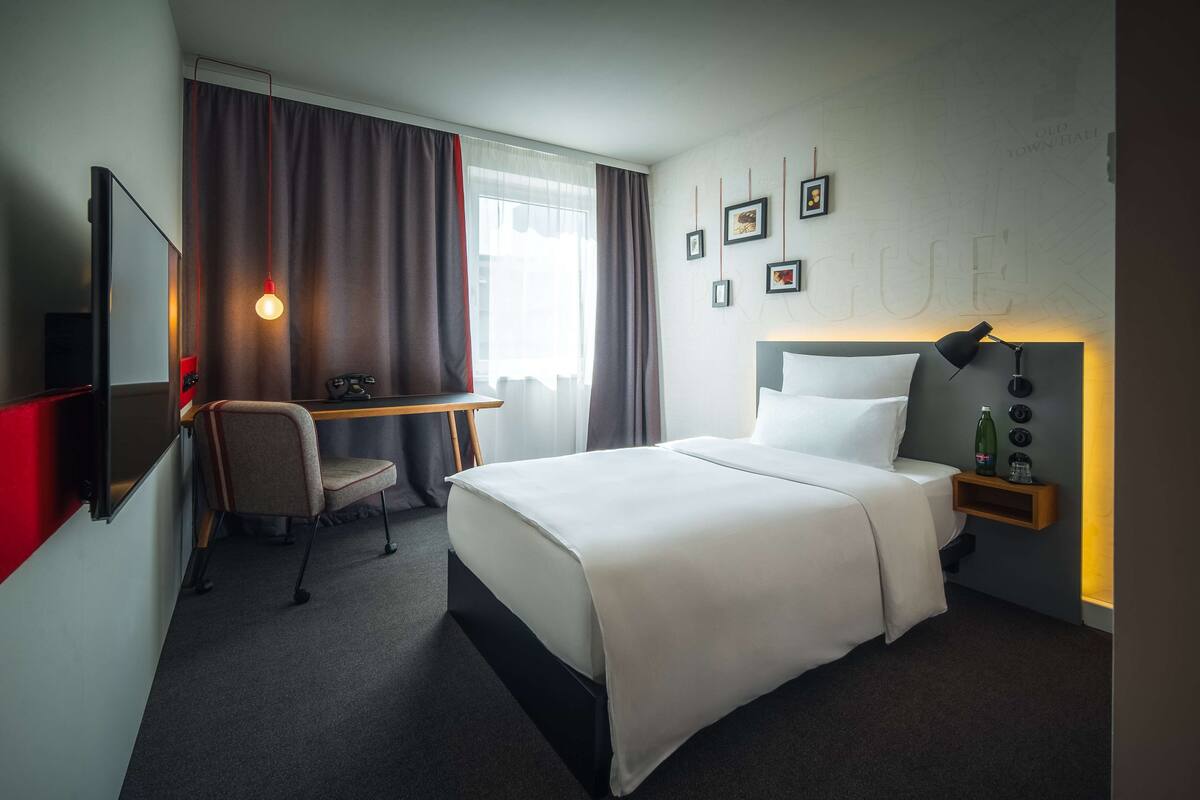 Habitación de hotel con cama, escritorio, silla, televisor y una pared con cuadros.