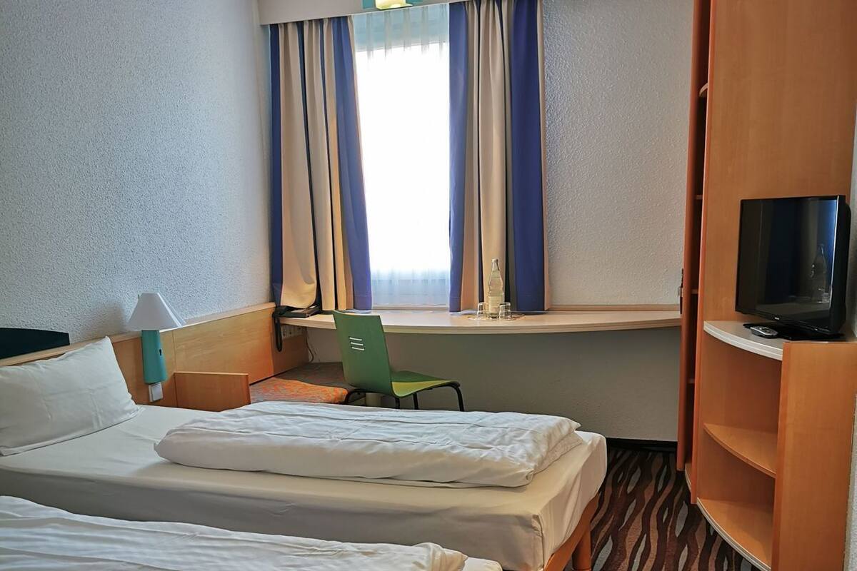 A hotel room with two beds, a TV, a chair, a window with curtains, and a patterned carpet.