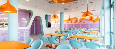 A brightly lit cafeteria with orange tables and blue chairs, featuring a modern design with pendant lighting.