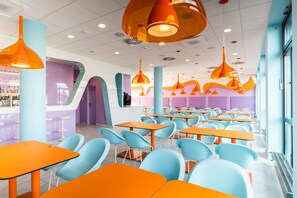 A brightly lit cafeteria with orange tables and blue chairs, featuring a modern design with pendant lighting.