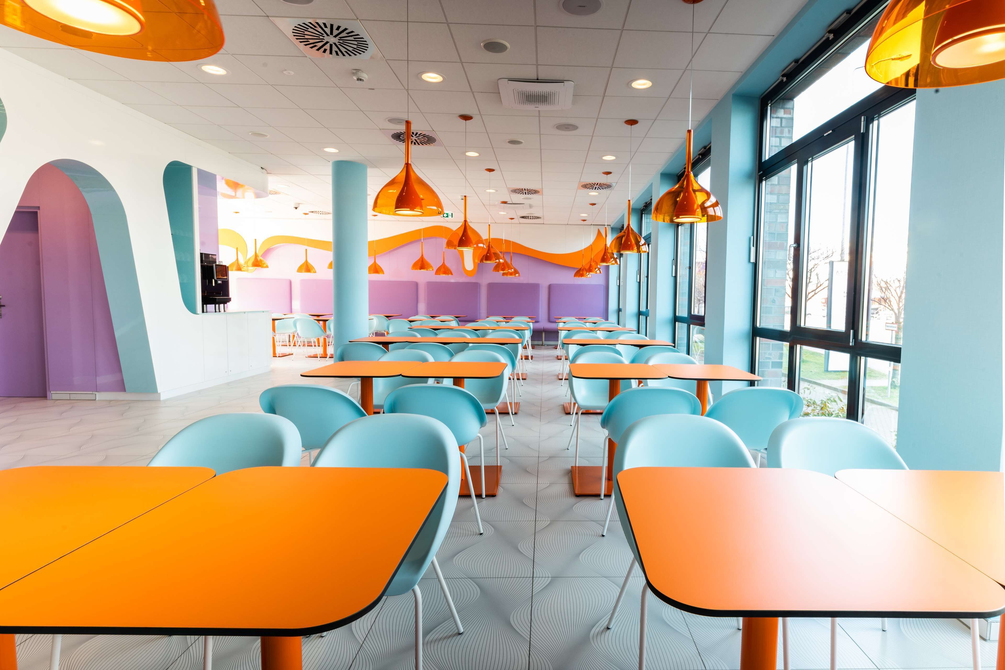 Un espace de restauration moderne avec des tables orange et des chaises bleues, de grandes fenêtres et des luminaires suspendus orange.