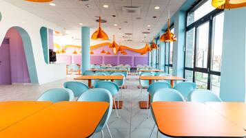 Un espace de restauration moderne avec des tables orange et des chaises bleues, de grandes fenêtres et des luminaires suspendus orange.