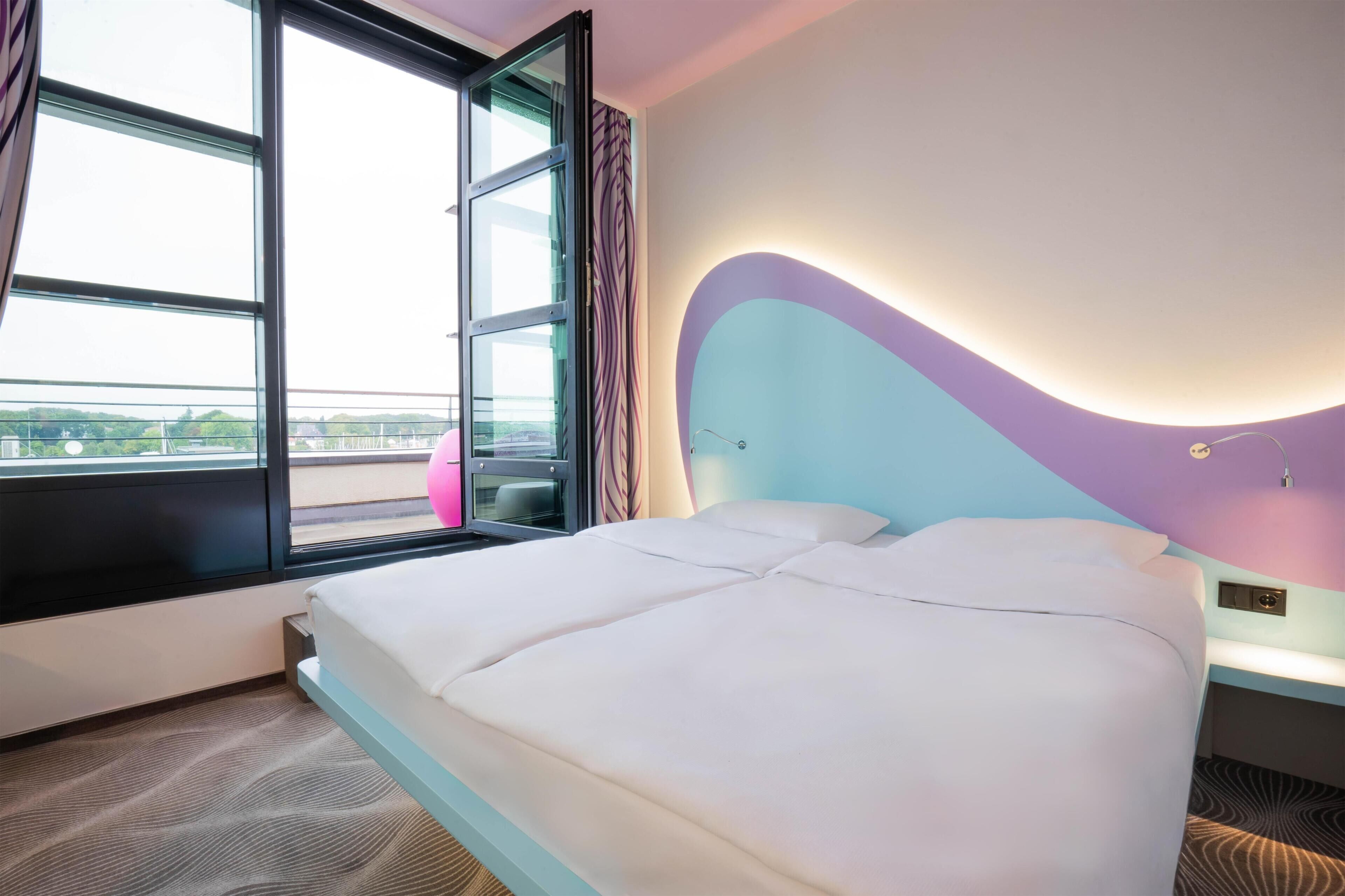 Habitación de hotel con cama, ventana, mesita de noche y vistas al exterior.