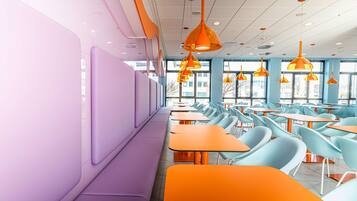 Un comedor moderno con asientos tipo banca color morado y mesas naranjas, ventanas amplias y lámparas colgantes naranjas.