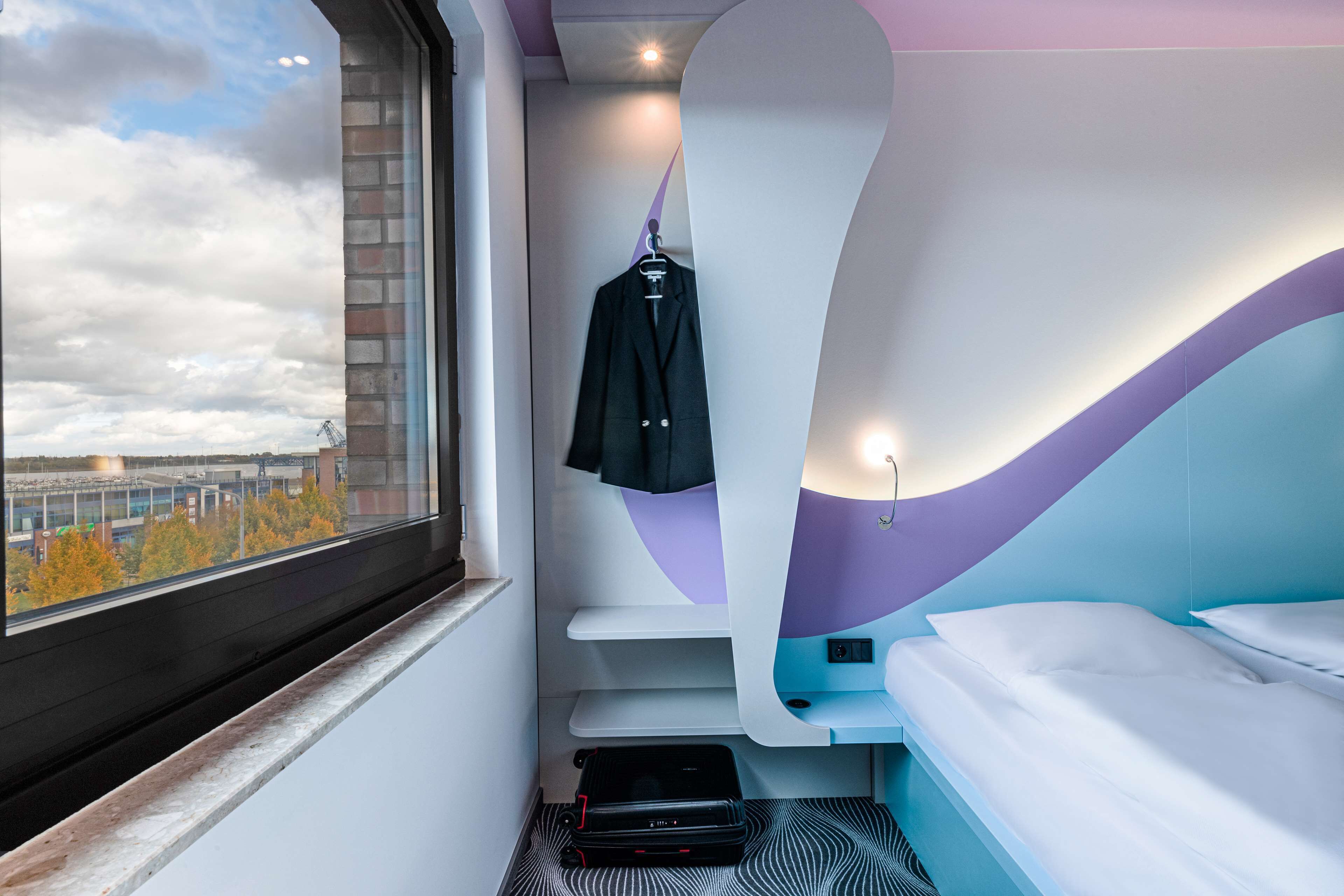 A modern hotel room with a large window, a bed, a desk, and a coat rack.