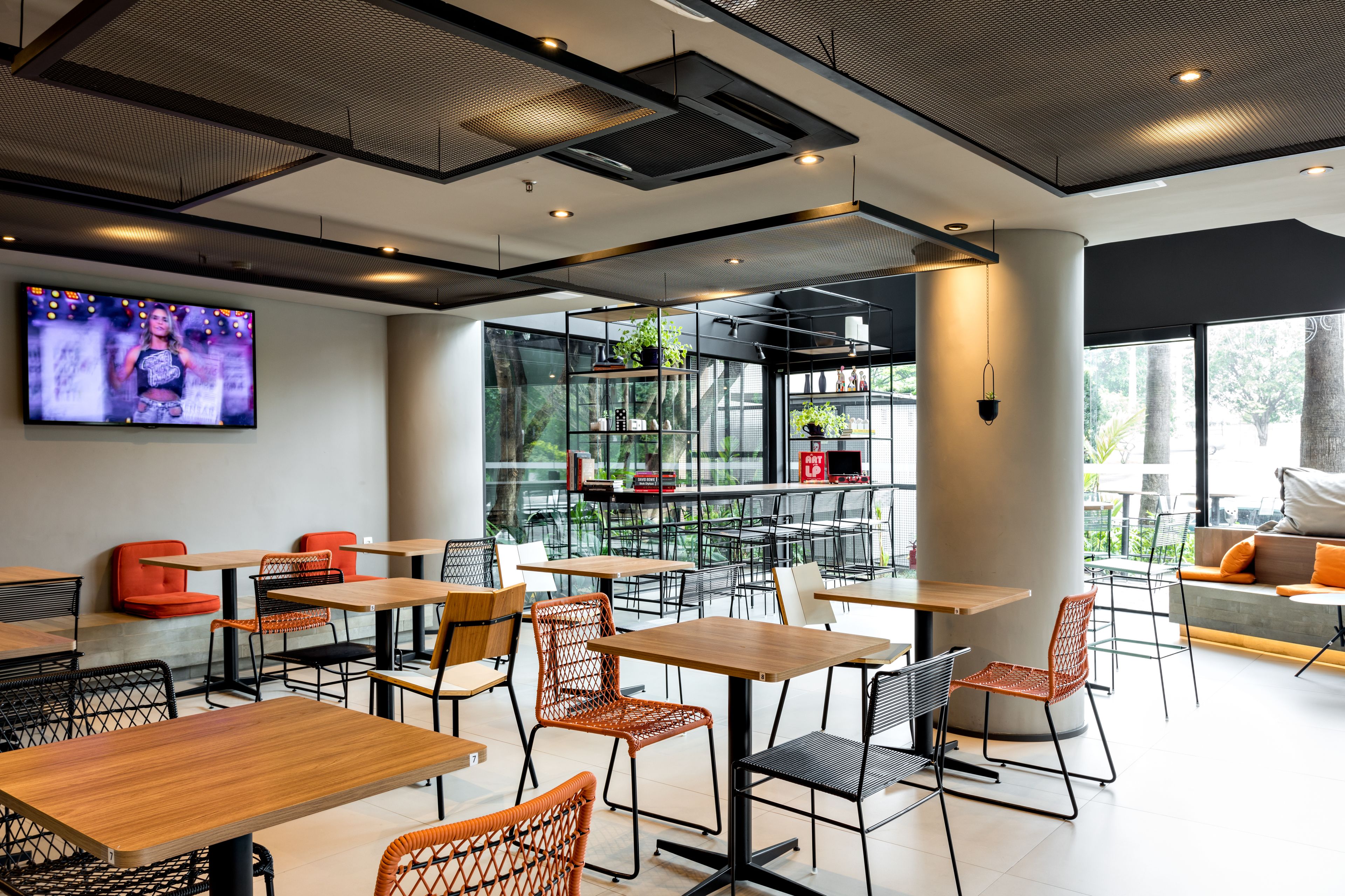 A modern cafe with wooden tables and chairs, a large TV screen, and a view of the outdoors.