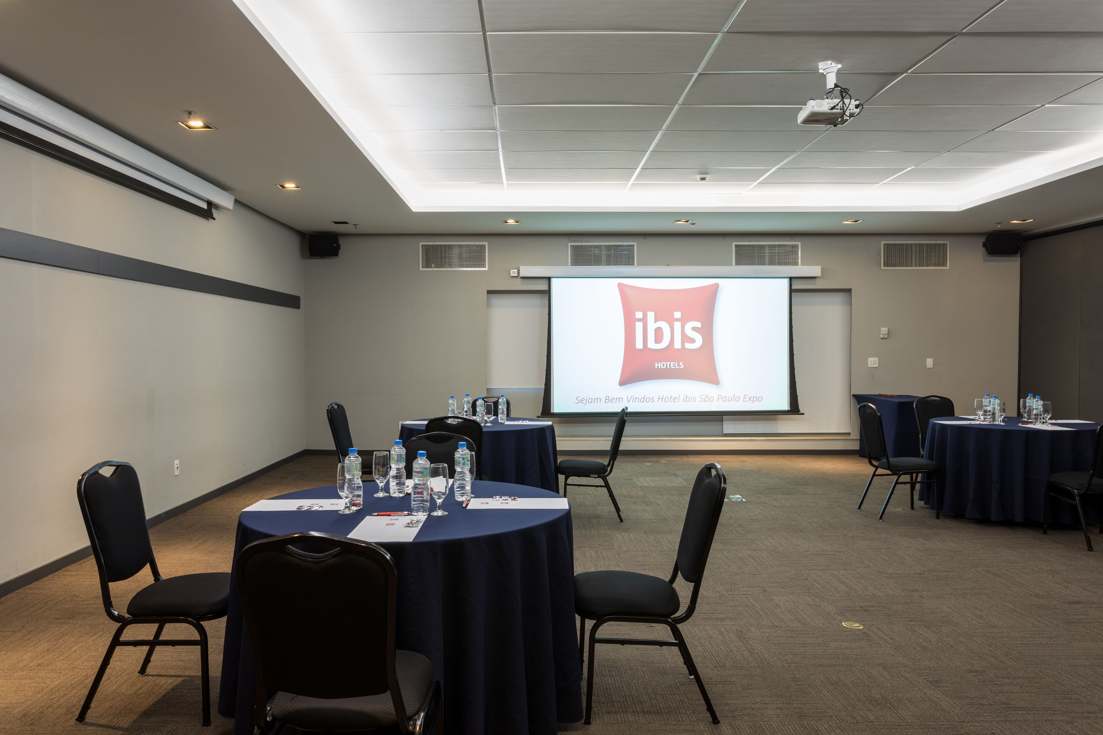 A conference room with round tables, chairs, and a projector screen displaying 'ibis Hotels'.