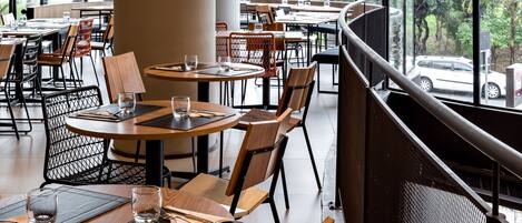 A modern restaurant with round tables, wooden chairs, and large windows offering a view of the outdoors.