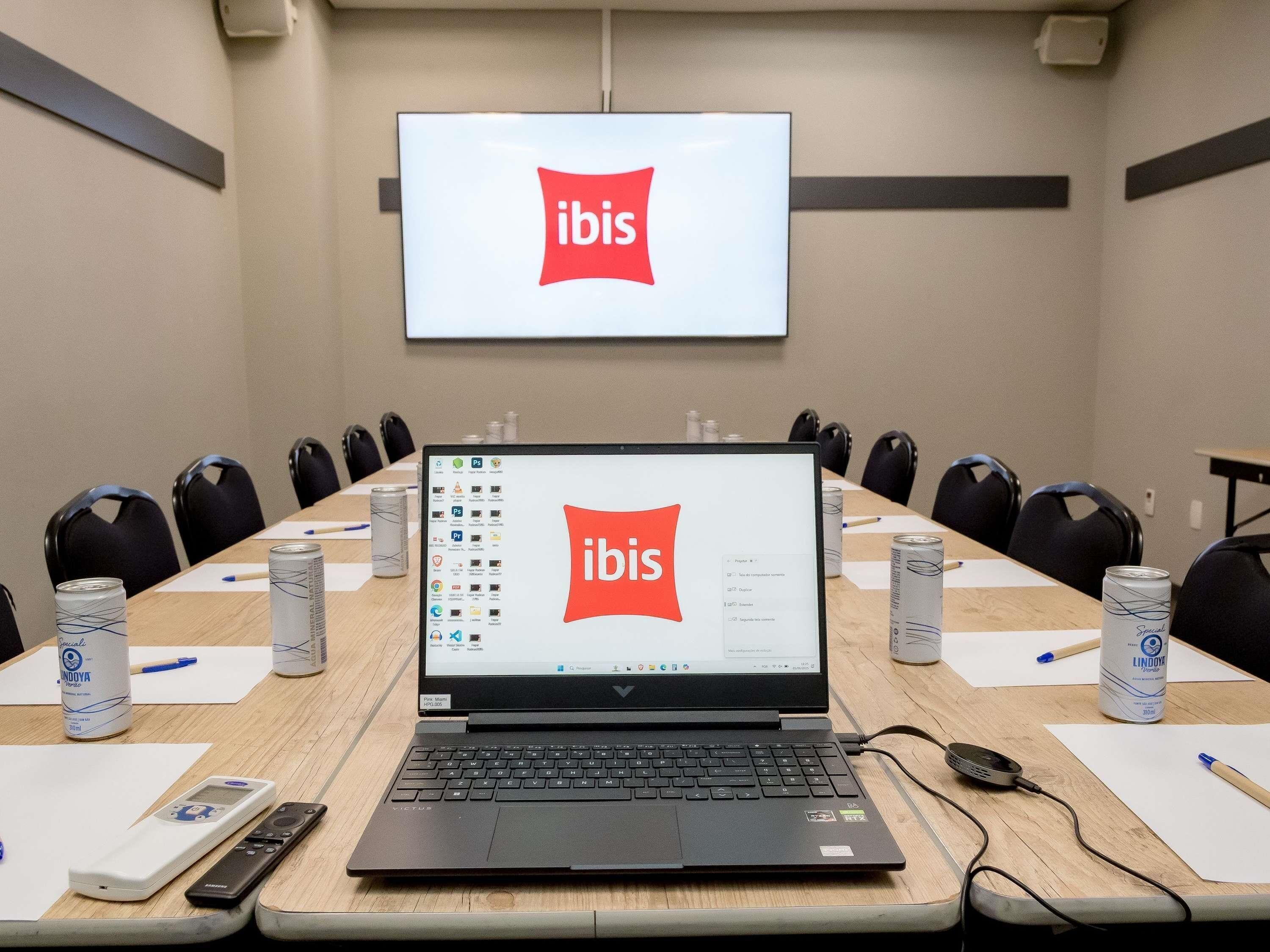 A conference room with a laptop displaying the ibis logo, a projector screen, and a table with chairs and cans of Lindoya beer.