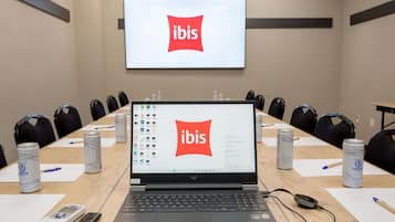 Uma sala de conferências com um laptop exibindo o logotipo da ibis, uma tela de projetor e uma mesa com cadeiras e latas de cerveja Lindoya.