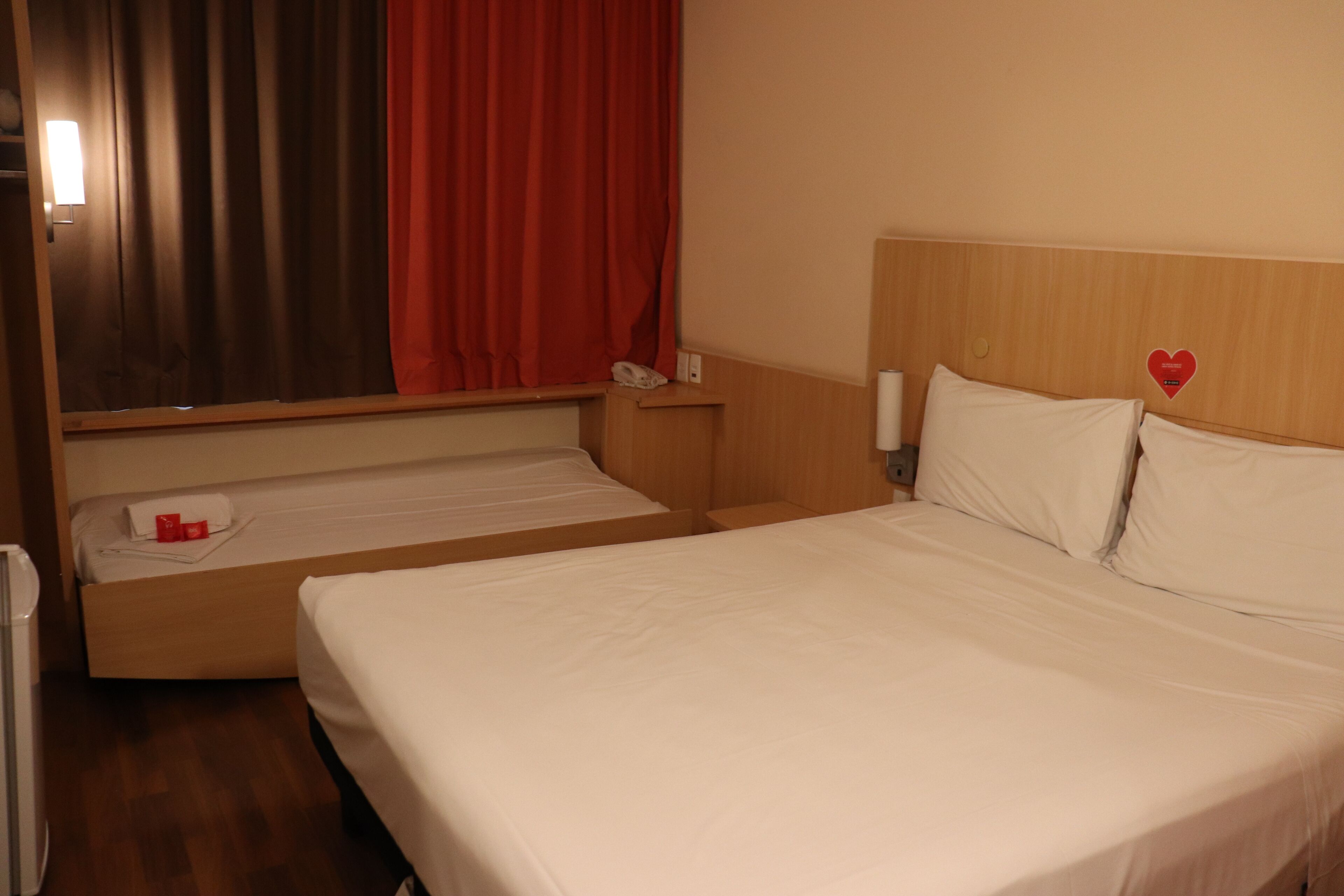 A hotel room with two beds, a wooden headboard, a red curtain, a bedside table with a phone, and a wall-mounted heart decoration.