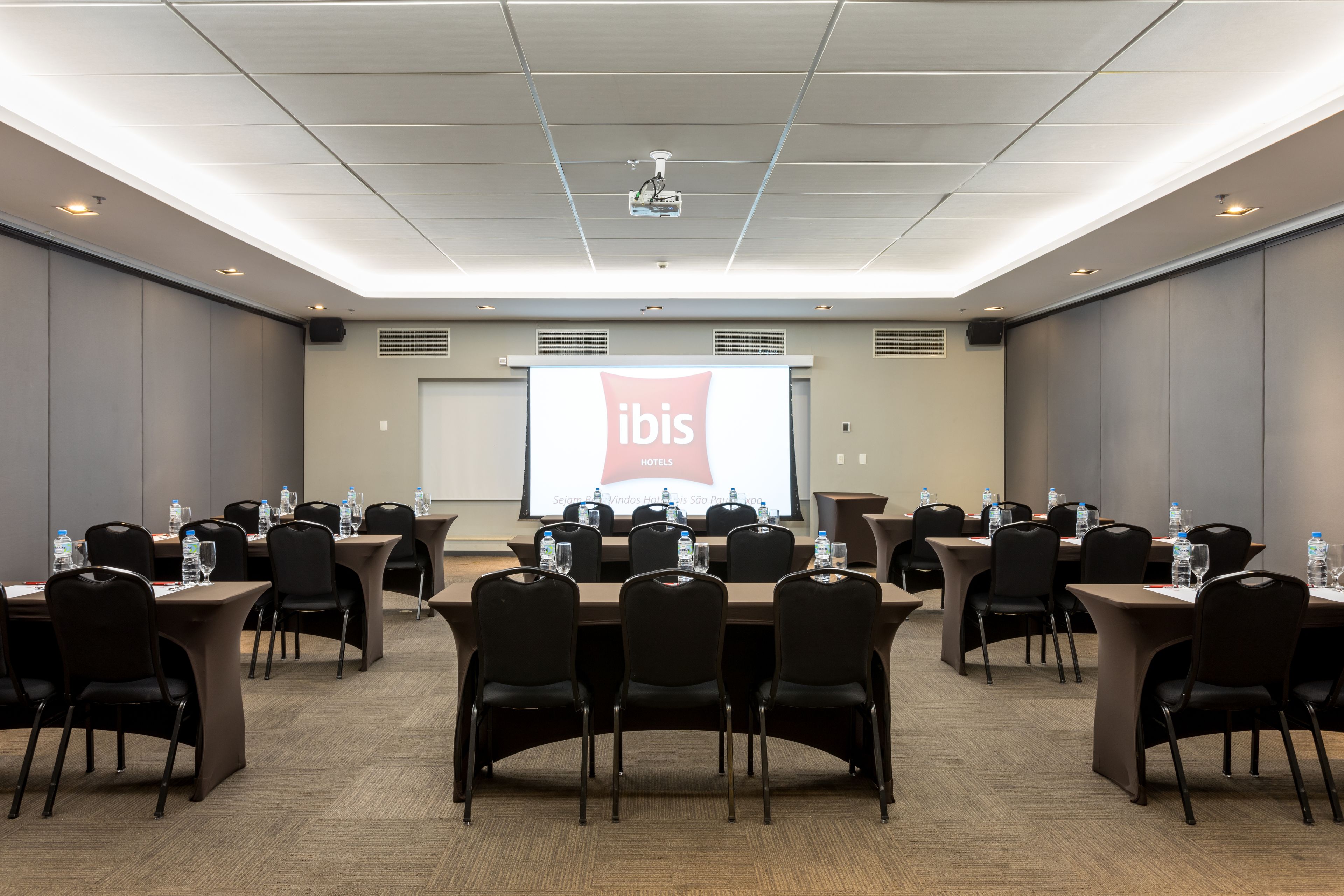 A conference room with a large screen displaying 'ibis HOTELS' and multiple tables with chairs and water bottles.