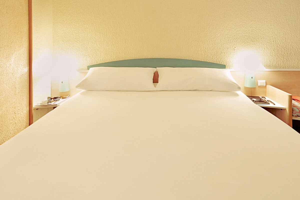 Een netjes opgemaakt bed met een groen hoofdeinde, twee nachtkastjes met lampen, en een houten nachtkastje met een lamp.