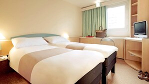 Une chambre d’hôtel avec un lit double, un bureau avec un ordinateur, une chaise, une fenêtre avec des rideaux et une paire de chaussures sur le sol.