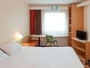 Ein Hotelzimmer mit einem Bett, einem Schreibtisch, einem Sessel, einem Fernseher und einem fensterbepflanzten Fenster.