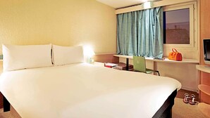 Une chambre d’hôtel avec un grand lit, un bureau avec une chaise, une fenêtre avec des rideaux, un téléviseur et une paire de chaussures posées sur le sol.