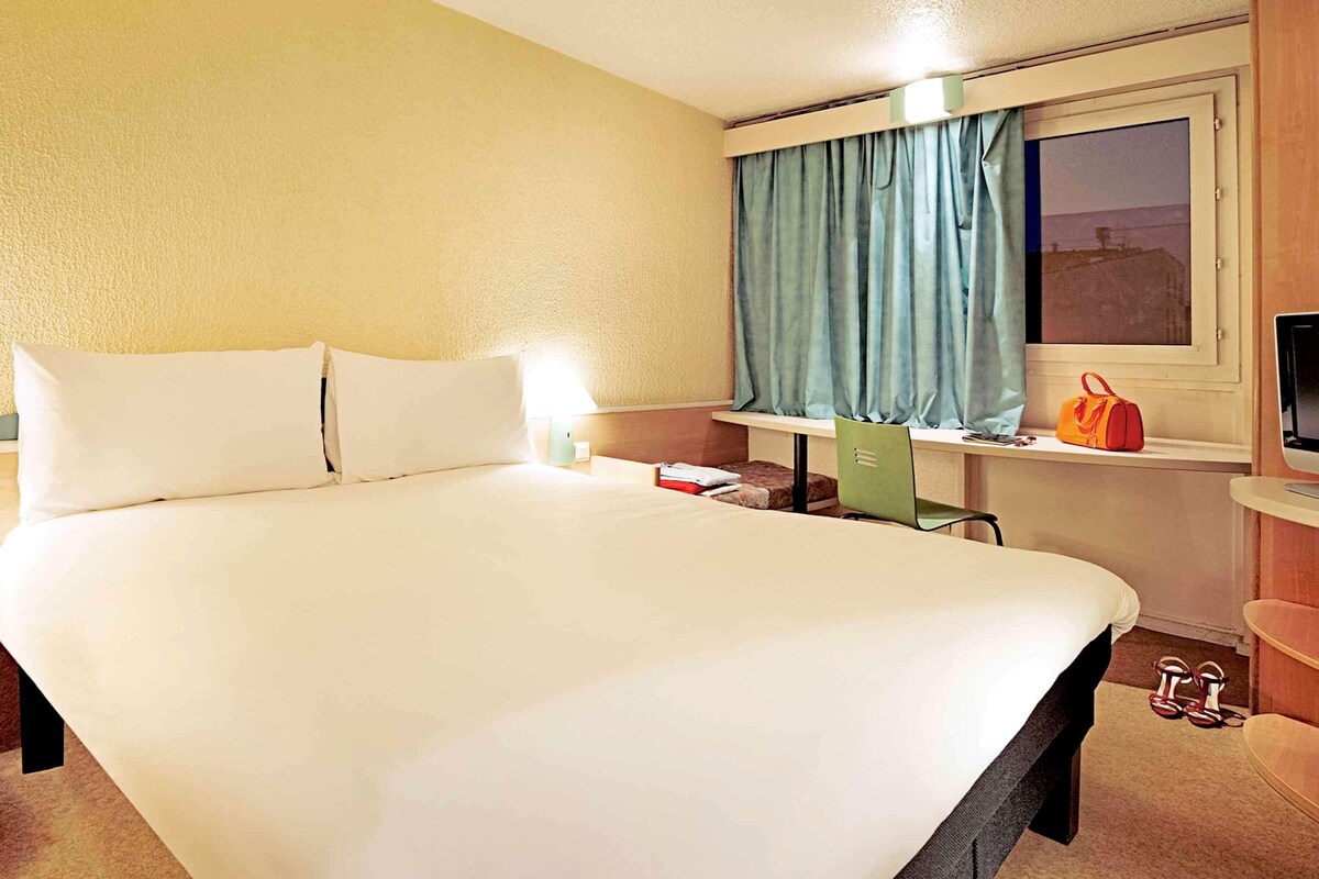 Een hotelkamer met een groot bed, een bureau met stoel, een raam met gordijnen, een televisie, en een paar schoenen op de vloer.