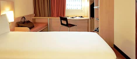A hotel room with a large bed, a desk, a chair, a lamp, and a wardrobe.