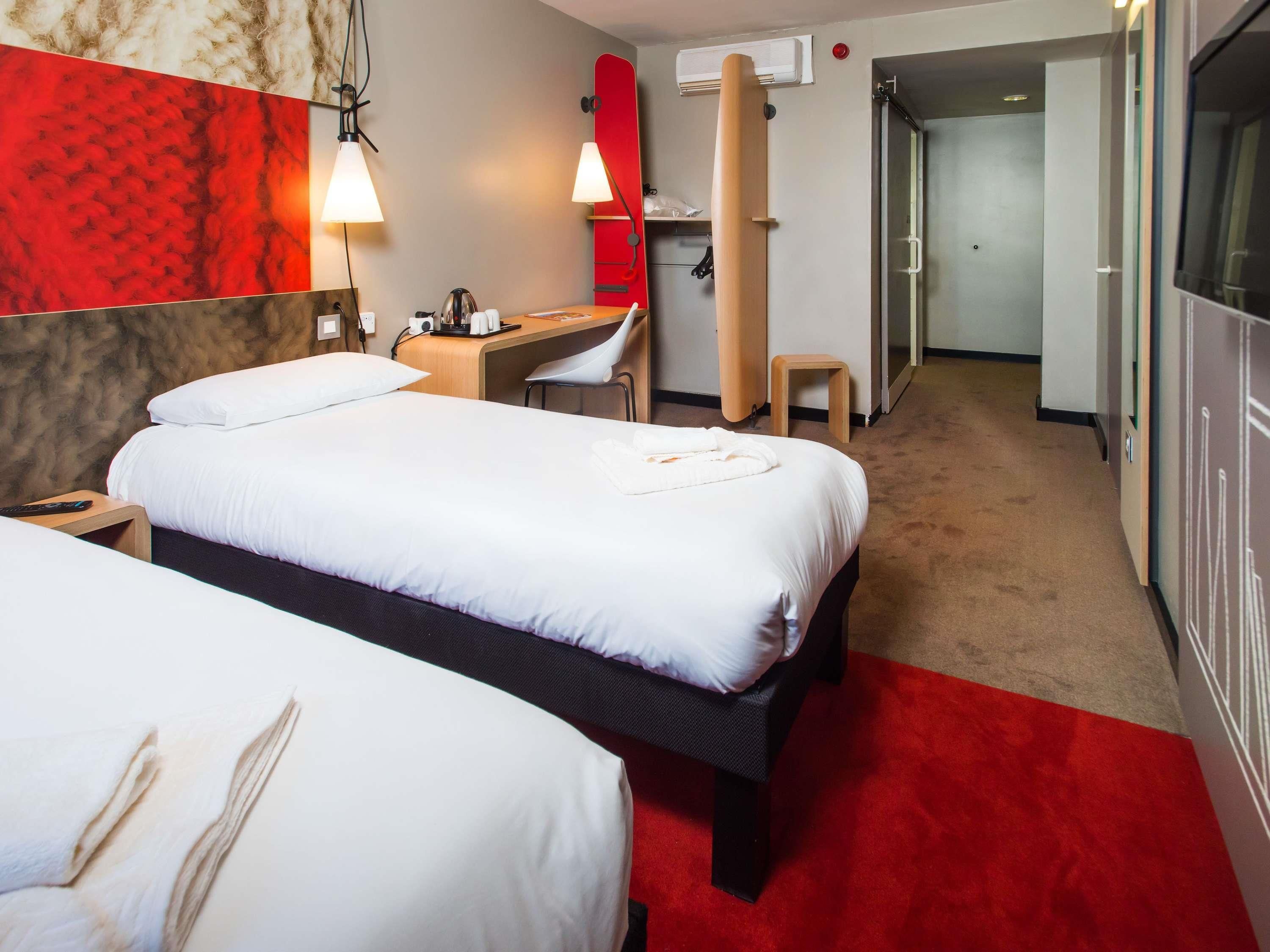 A hotel room with two beds, a desk, a chair, a red wall, and a hallway leading to a bathroom.