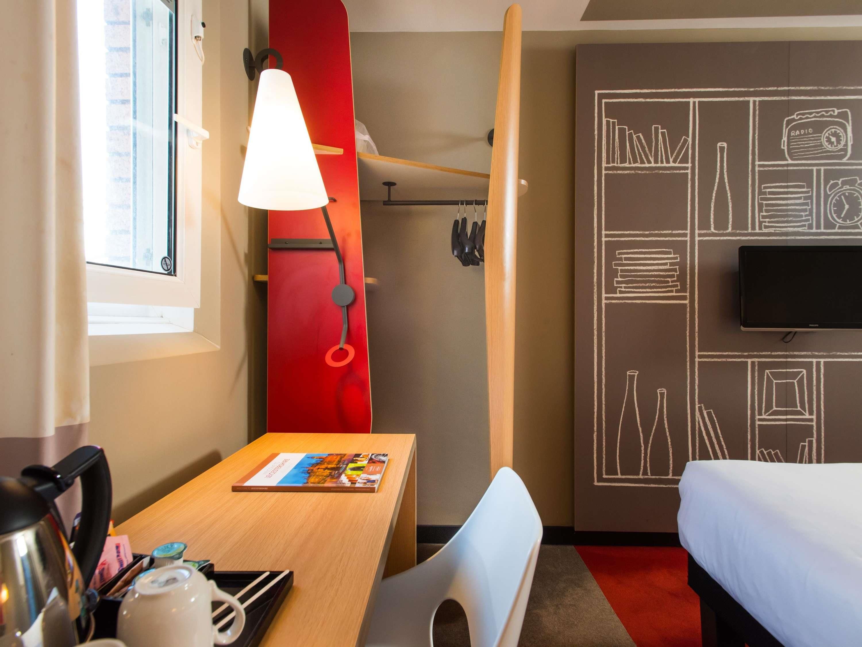 A hotel room with a wooden desk, a red wall with white drawings, a TV, a bed, and a window with curtains.