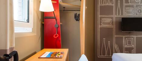A hotel room with a wooden desk, a red wall with white drawings, a TV, a bed, and a window with curtains.