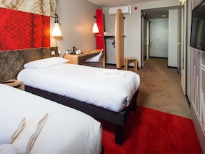 A hotel room with two beds, a desk, a chair, a red wall, and a hallway leading to a bathroom. - ibis Sheffield City (Sheffield)